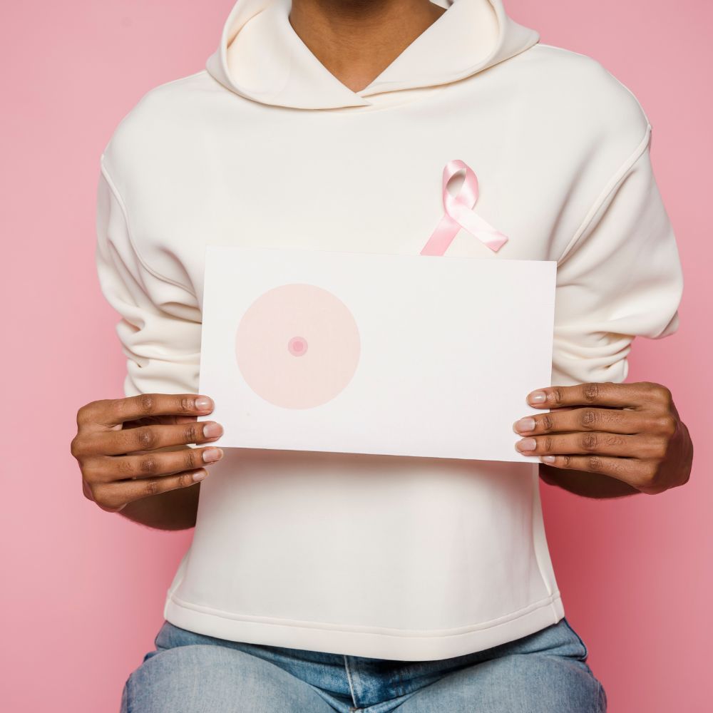 Person in a white hoodie with a pink ribbon on the chest, holding a white card with a pale pink circle design against a pink background (breast cancer awareness)