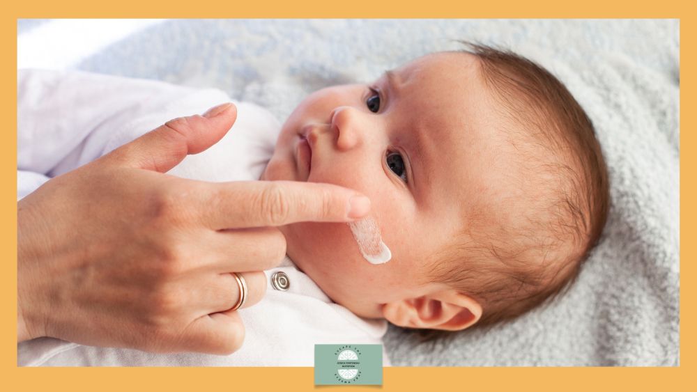 Caregiver applies a small amount of cream to a baby's cheek while the baby lies on a soft blanket